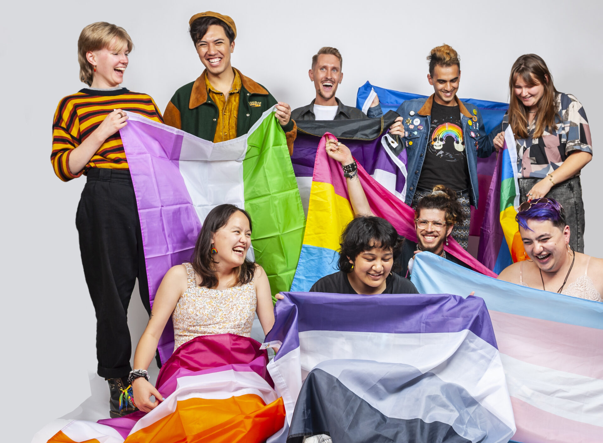 Inside OUT youth volunteers. Studio portrait photograph of a diverse group of people holding various pride flags against a plain background.