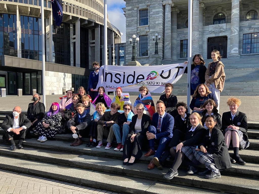 Inside OUT at Parliament. A diverse group of people carrying an Inside OUT banner seated on the steps before the New Zealand Parliament "Beehive" building.