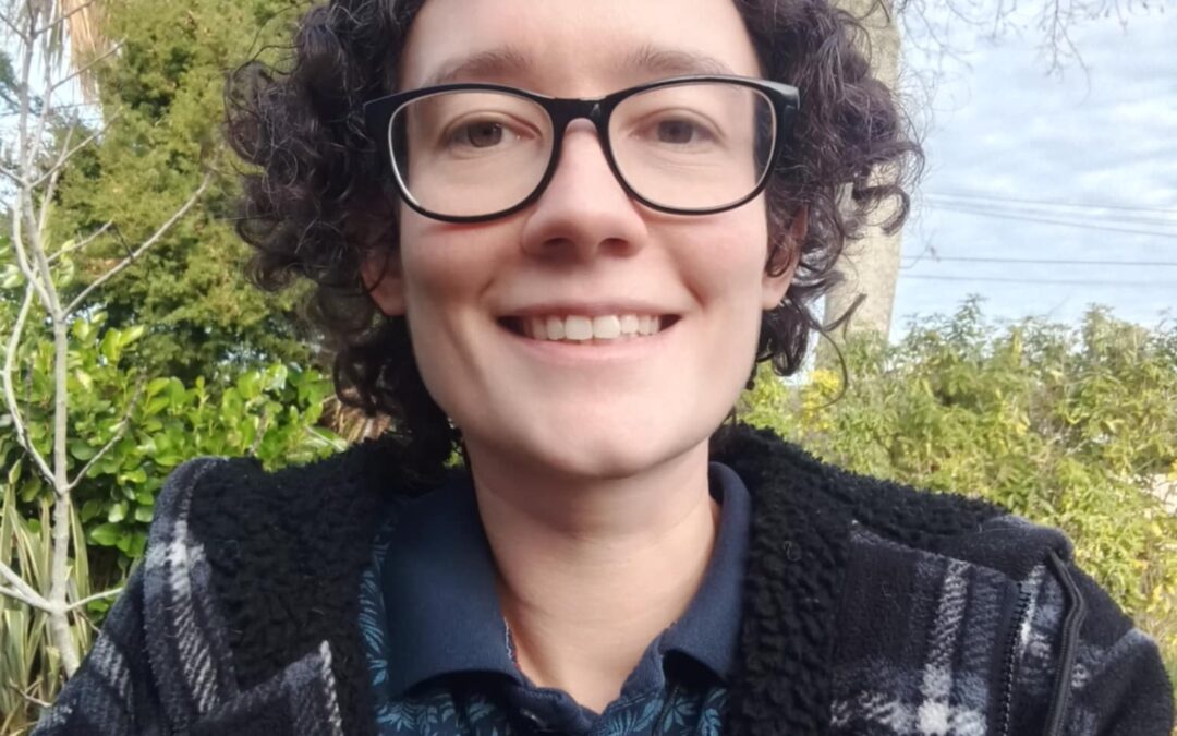 A person with light skin, short dark curly hair and glasses smiling.