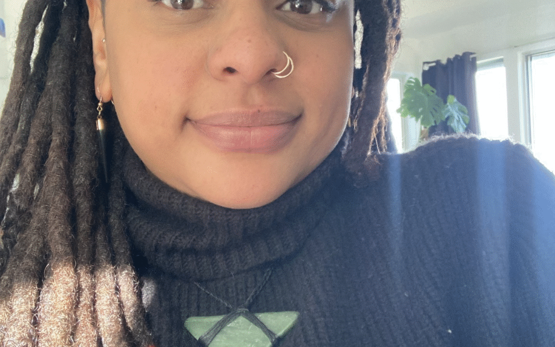 Headshot of a person smiling with brown skin and locs wearing a black turtleneck and pounamu.