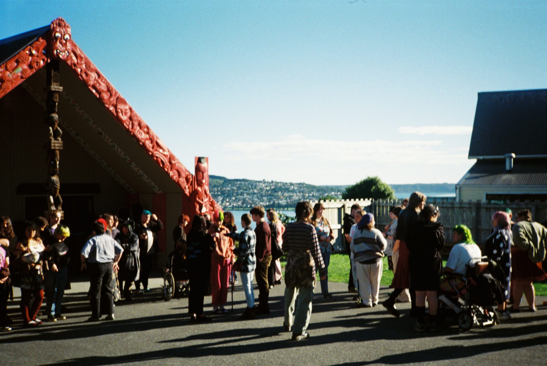 A range of young people captured in a candid photo beside a wharenui with the ocean in the background and a blue sky. The image is slightly out of focus so there is a slightly blurred effect on the photo.
